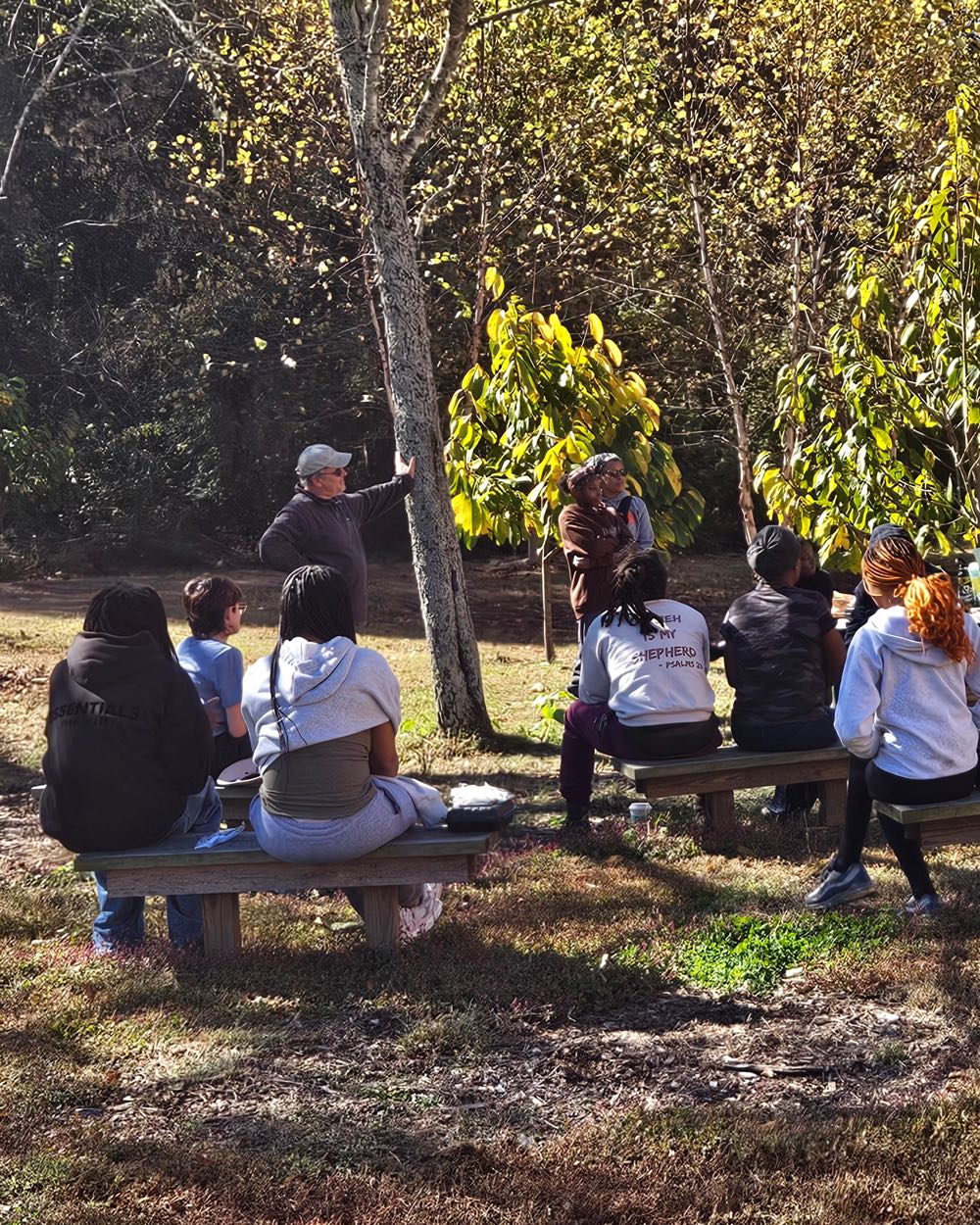 A class seated on benches under the trees at the edge of the farm, listening to a talk