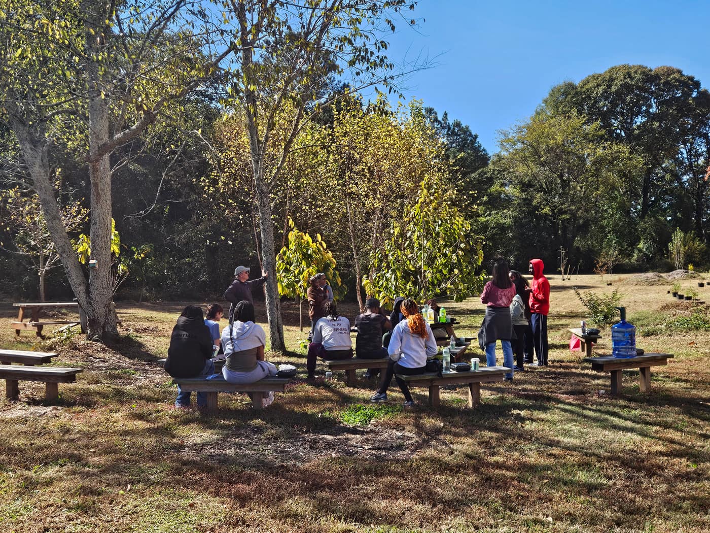 A class seated on benches under the trees at the edge of the farm